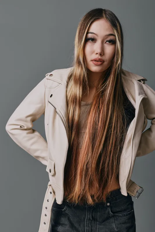 Young woman with long straight hair wearing a light jacket and dark pants stands with hands on hips against gray background looking confident and stylish.