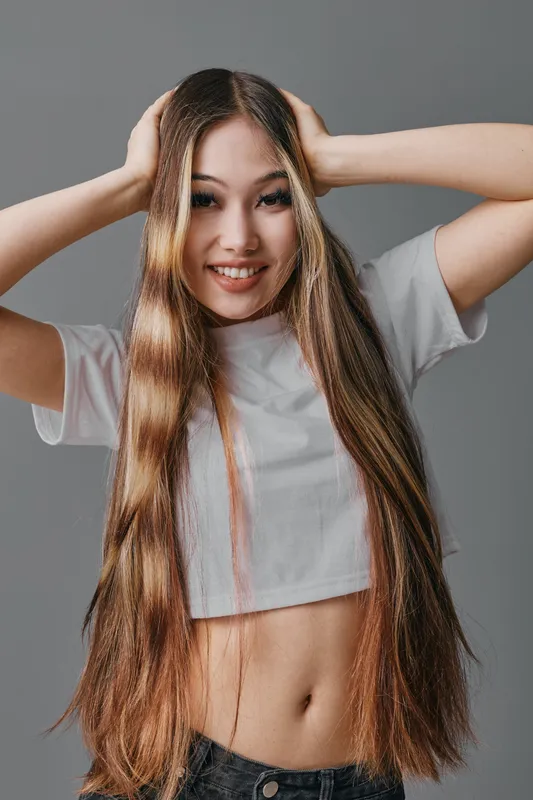 Young woman with long straight hair smiling and posing with hands on head wearing cropped white t shirt and dark pants on gray background indoors.