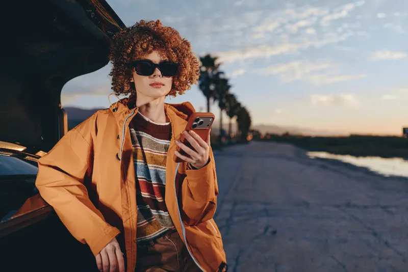 Woman lifestyle portrait with curly hair, sunglasses, and smartphone in hand, standing by a parked car on the road at sunset in golden evening light, coastal travel mood.