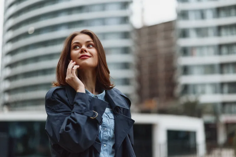 Woman stands outdoors in an urban setting, talking on a mobile phone, wearing a dark coat over a denim shirt, with a thoughtful expression and modern city backdrop.