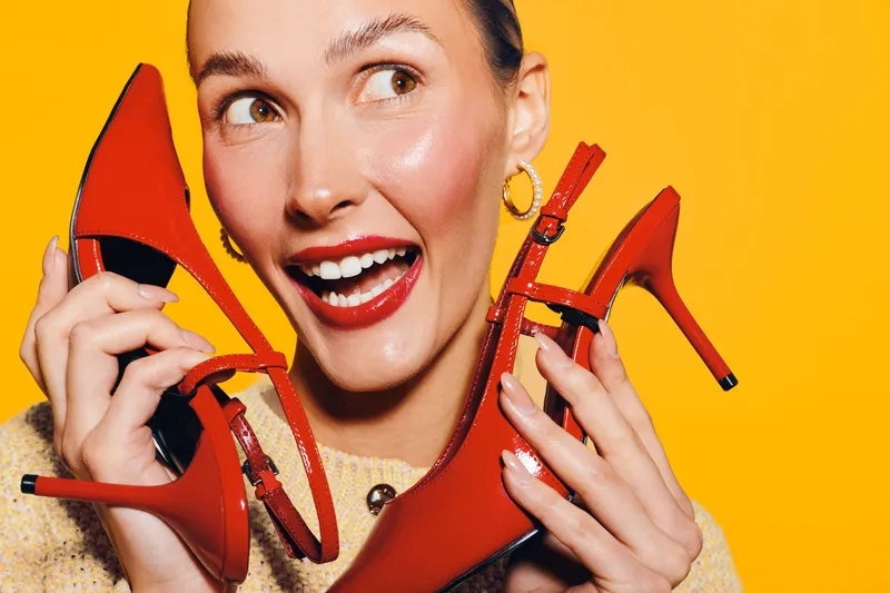 Happy woman with red lipstick holding red high heel shoes near her face on yellow background. Concept of fashion, style, shopping, and joyful emotions in studio lighting.