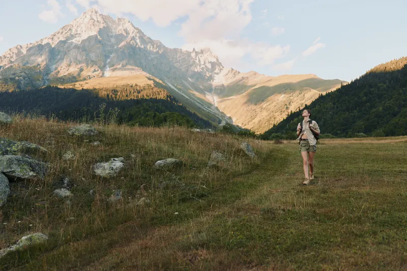 Woman hiker walks across a grassy valley with towering mountains in the distance, enjoying outdoor travel, nature, and peaceful solitude, backpack glinting in warm sunlight as grasses ripple