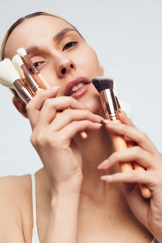 A close-up portrait of a young woman holding various makeup brushes near her face with a neutral expression against a plain background. Concept for beauty and cosmetics.