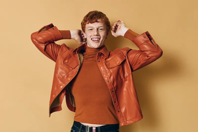Young man smiling with red hair and freckles wearing brown leather jacket and turtleneck sweater posing against beige background with hands behind head looking confident and happy.