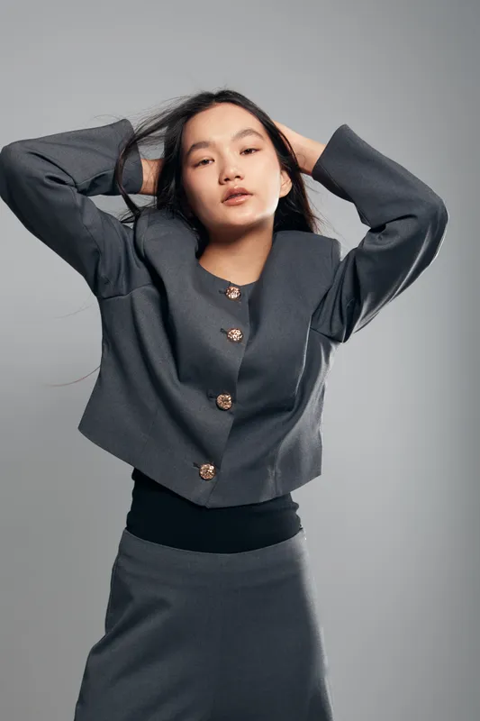 Woman posing with hands in hair wearing stylish gray jacket and skirt on neutral background. Fashion model portrait with confident expression indoors for clothing catalog or style.