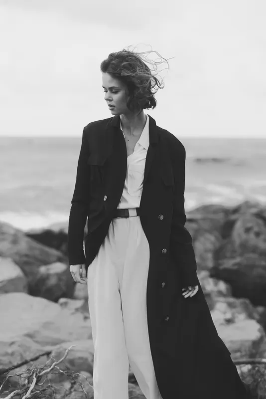 Fashion, coat, wind, ocean, woman on rocky shore in monochrome, long coat and light suit, wind swept hair, moody seaside scene highlighting style and elegance