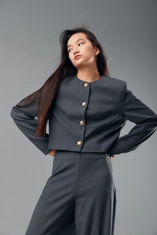 Young woman wearing fashionable dark gray suit with long hair standing confidently posing against a plain light gray background studio portrait fashion style.