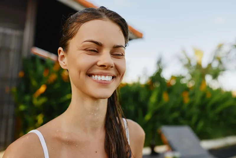 Happy outdoor portrait of a smiling woman enjoying a carefree moment in sunlight, casual summer vibe, serene garden background, natural makeup, relaxed posture and warm mood, inviting lifestyle scene