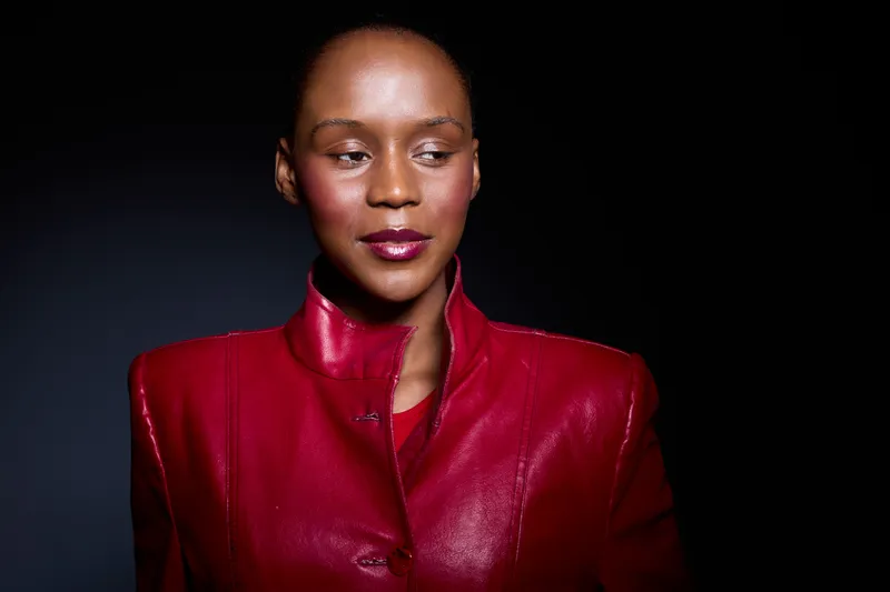 Elegant black woman with dark skin wearing a red leather jacket poses confidently against a dark background. Studio portrait highlighting fashion and beauty concepts.