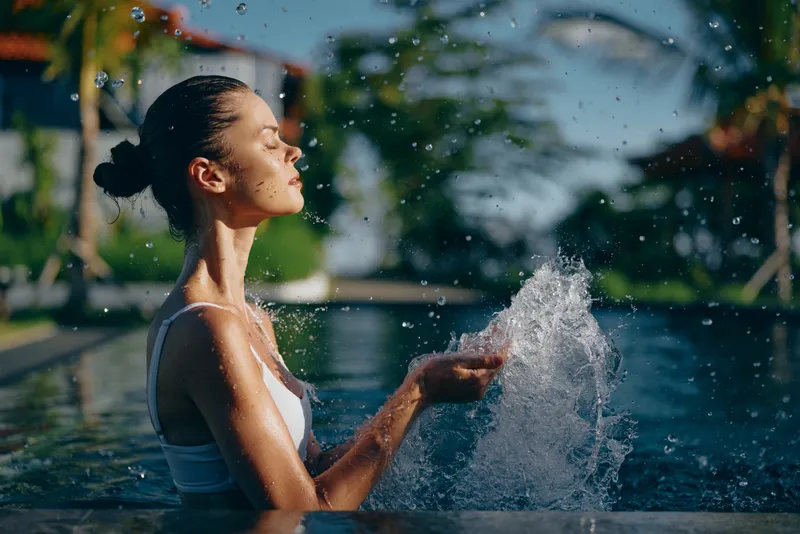 Young woman in white swimsuit enjoying water splash in outdoor pool with eyes closed. Relaxation and lifestyle concept with natural sunlight and tropical background.