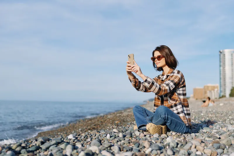 Woman sitting on a pebbled beach taking a selfie with her phone, wearing a plaid shirt and jeans, sunglasses, enjoying sunny coastal scenery and calm ocean waves.