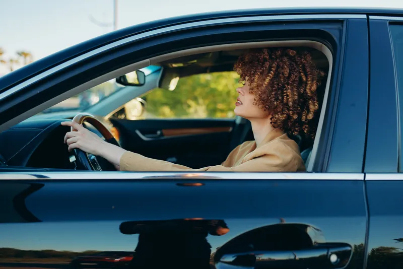 Vibrant woman driving a car, enjoying a sunlit moment with relaxed posture, soft breeze in hair, retro film color adds warm tones to a candid lifestyle scene in a bright background scene