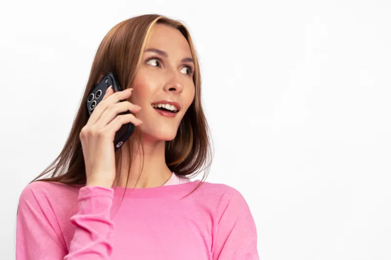Young woman in pink sweater talking on smartphone looks aside with surprised expression. Isolated on white background. Concept for communication, technology, mobile phone, and emotions.