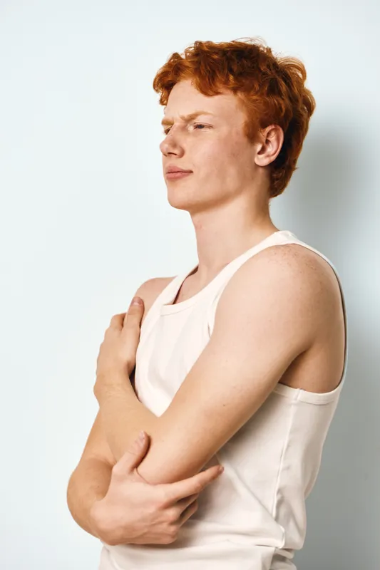 Young redhead man in white sleeveless shirt with crossed arms looking thoughtfully aside on white background. Casual style, confident expression and relaxed posture portrait.