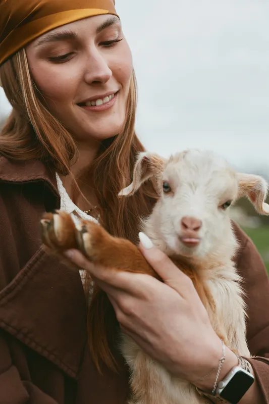 Smiling woman wearing a brown coat holds a young goat kid in an outdoor setting, close-up of a gentle moment. Lifestyle scene highlighting animal bond, care, and nature. Warm light creates calm,
