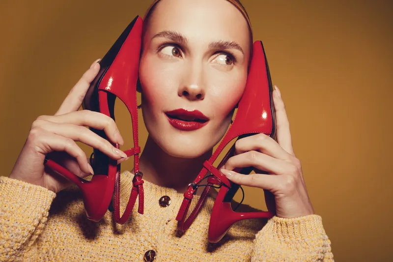 Woman with red lipstick holds red high heel shoes near face, wearing yellow knitted sweater. Studio portrait on brown background. Concept of fashion, style, and beauty accessories.