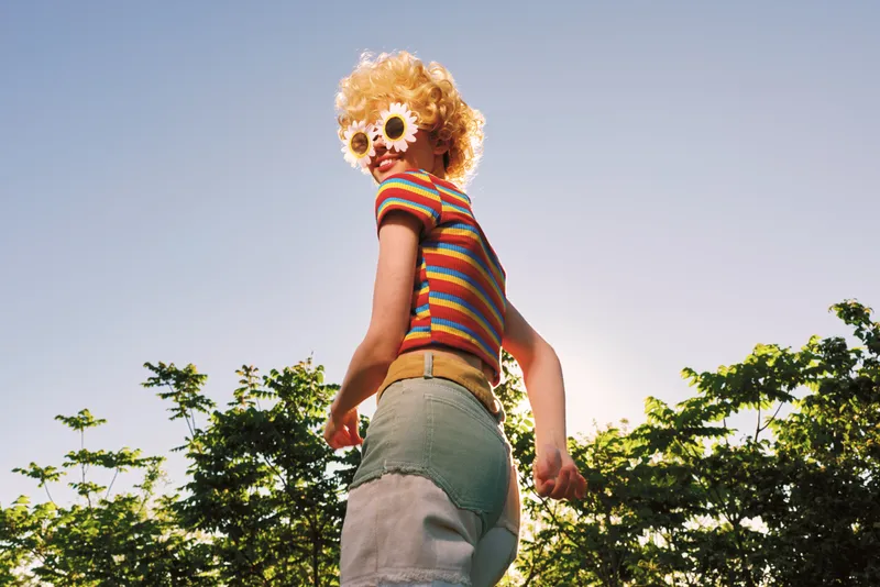 Retro vintage lifestyle woman outdoors, wearing striped shirt and denim shorts, with flower sunglasses, posing playfully under bright sunshine.
