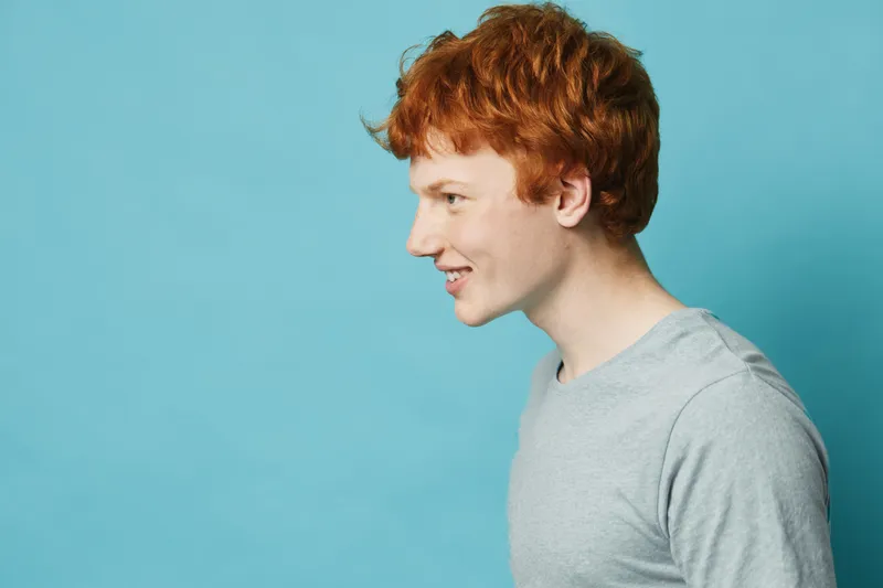 Profile of young man with red hair smiling in grey shirt on blue background. Side view portrait of happy male person with casual hairstyle and light skin in studio.