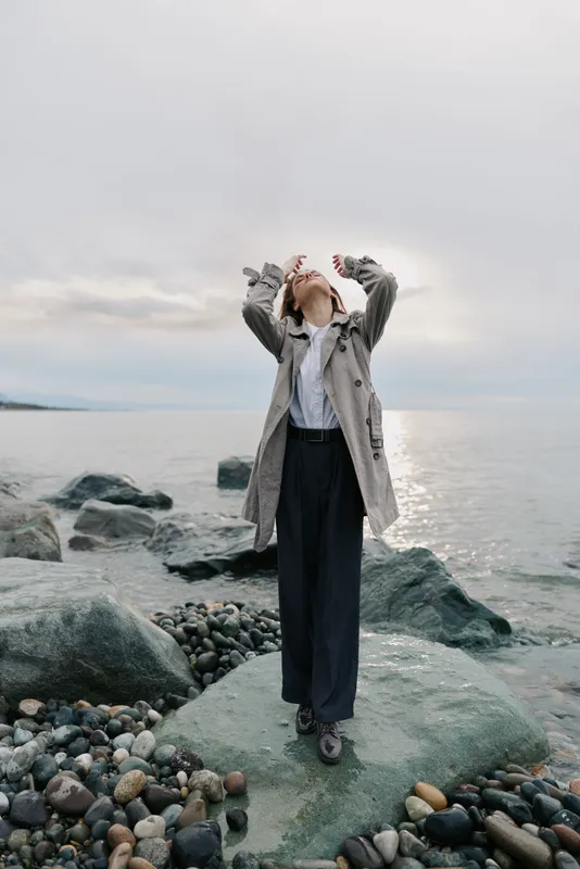 Woman in patterned coat stands on rocky shore looking up with hands raised. Cloudy sky and calm sea in background create peaceful and contemplative mood by the water.