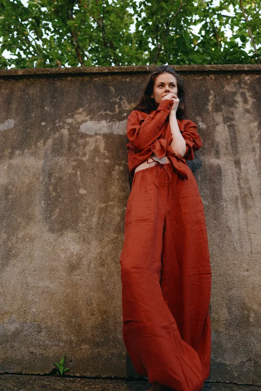 Young woman in a crimson dress stands against a weathered wall, surprised expression visible as she covers her mouth with her hands, outdoors with green foliage in the background