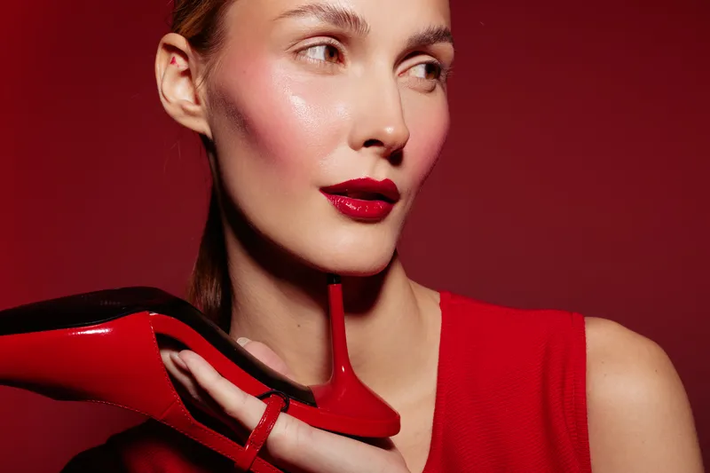 Close-up of a stylish woman with red lipstick and red dress holding a red high heel shoe near her face. Concept of fashion, beauty, elegance, and glamour in studio lighting.