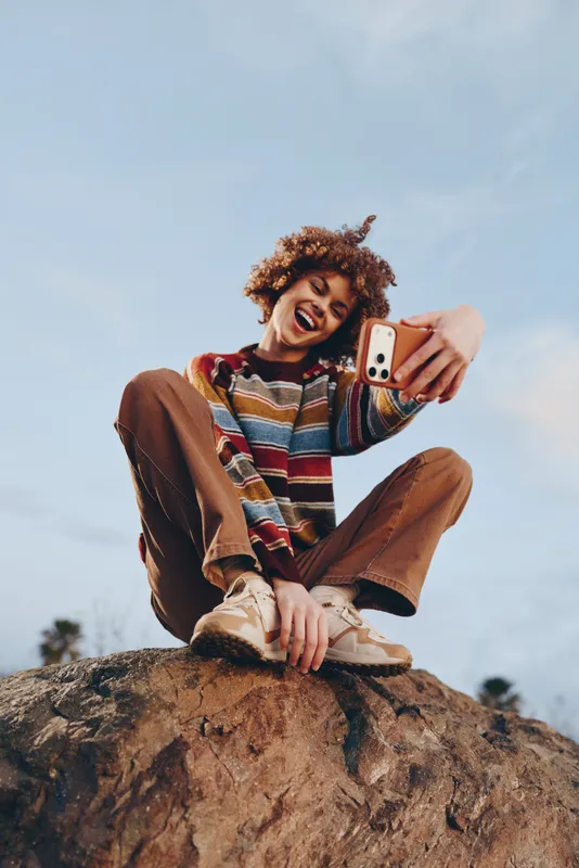 Cheerful woman lifestyle outdoors, sitting on a rock, smiling and laughing while holding a smartphone for a selfie, curly hair in the sun, natural colors, blue sky, fresh breeze today