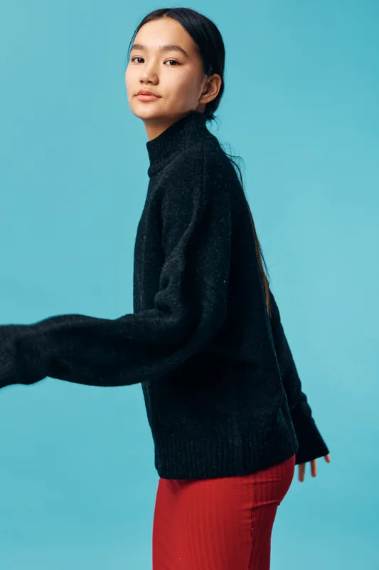 Young woman in black sweater and red skirt stands in front of blue background, looking confidently with relaxed posture and minimal makeup for fashion portrait.