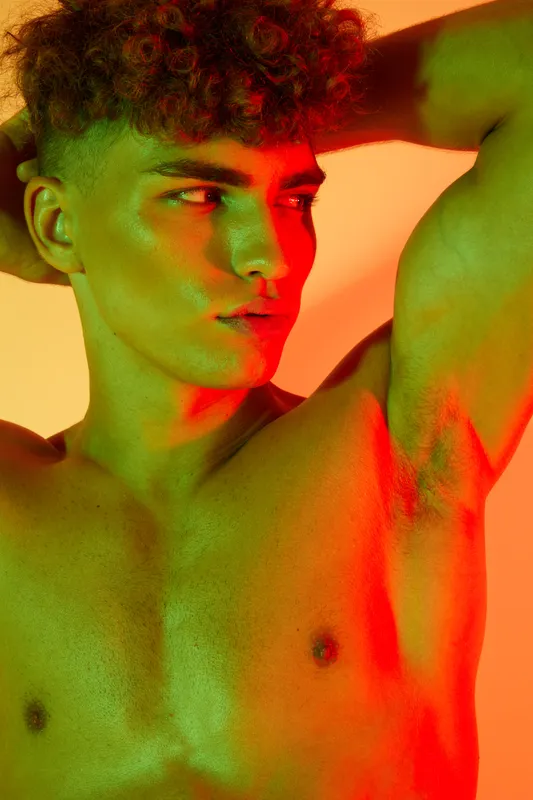 Young man with curly hair posing shirtless with hands behind head, colorful lighting highlighting his face and upper body in studio portrait with orange and green tones.