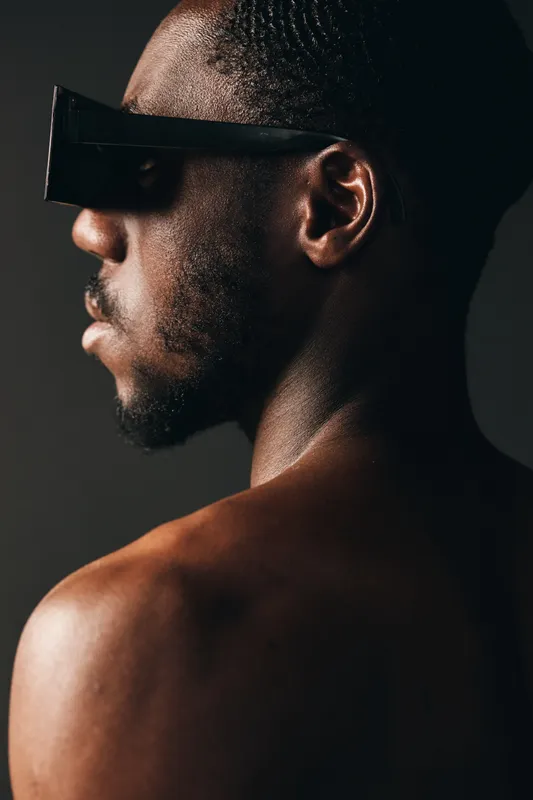 African man with dark skin wearing futuristic sunglasses in creative conceptual visual style, closeup portrait with bare shoulders and textured skin on dark background.