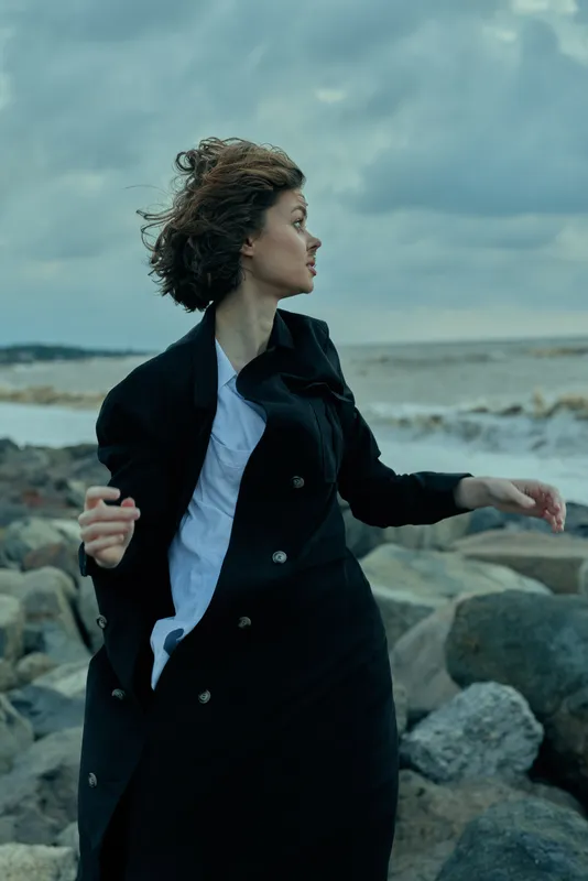 Woman on a rocky coast facing strong wind in a dark coat, sea spray in the air, moody sky, motion and outdoor style, dramatic breeze, ocean backdrop, candid atmosphere