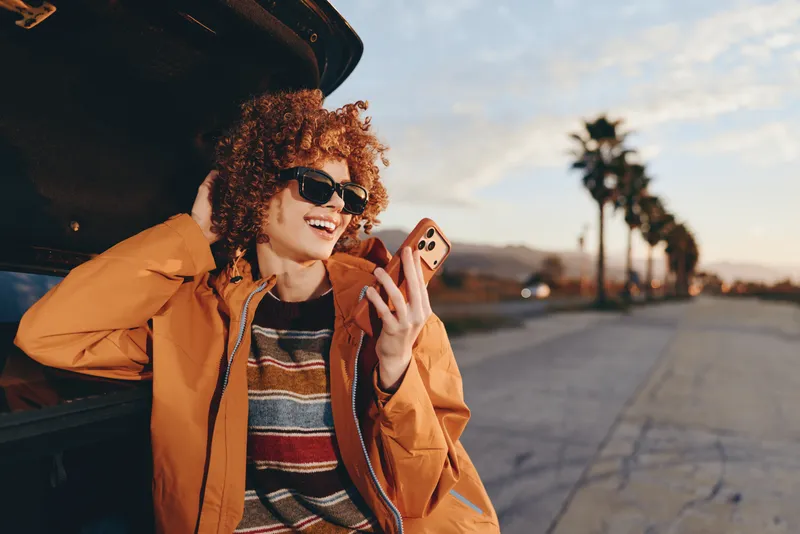 Woman lifestyle portrait outdoors at sunset, curly hair sunglasses, wearing orange jacket, smiling while holding phone and looking forward, warm sky and city road create relaxed travel mood
