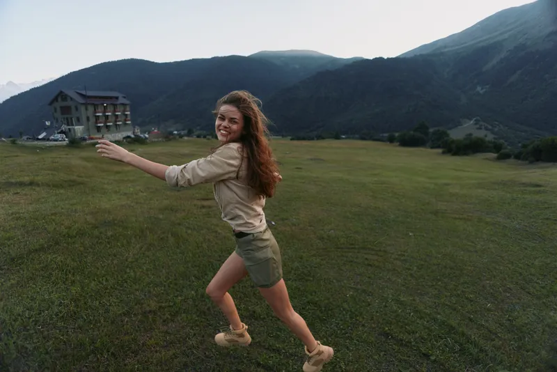 Woman lifestyle shot of a young woman running across a green field with distant mountains, smiling and enjoying outdoor freedom in bright daylight.