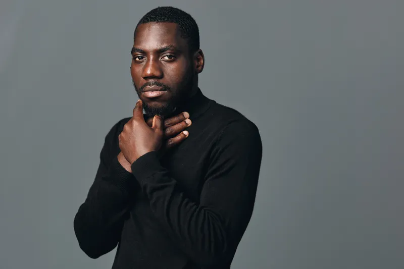 African man with dark skin wearing black sweater posing with hand on chin in creative conceptual visual style studio portrait on gray background for fashion and confidence.