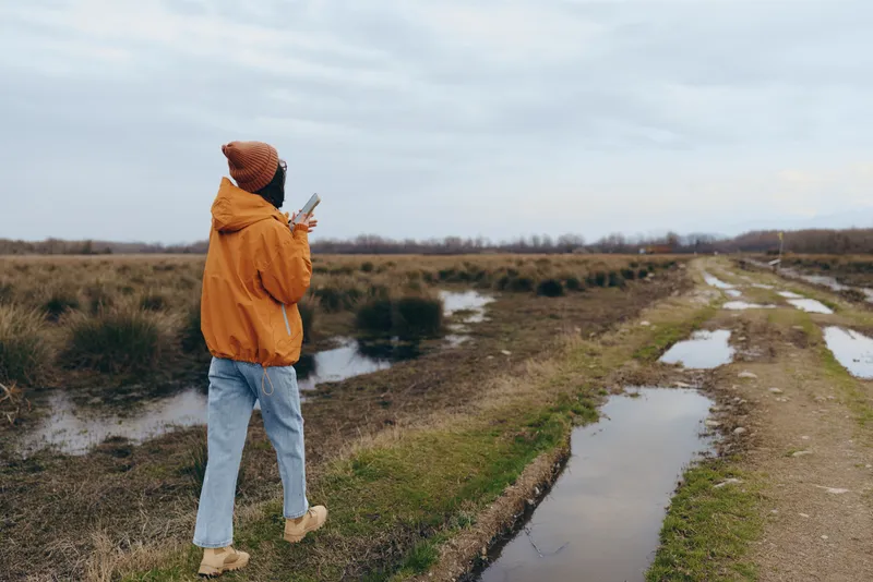 Woman outdoors lifestyle, walking on a dirt path across a grassy field, facing the horizon, cloudy sky, puddles of water beside the trail, calm quiet mood
