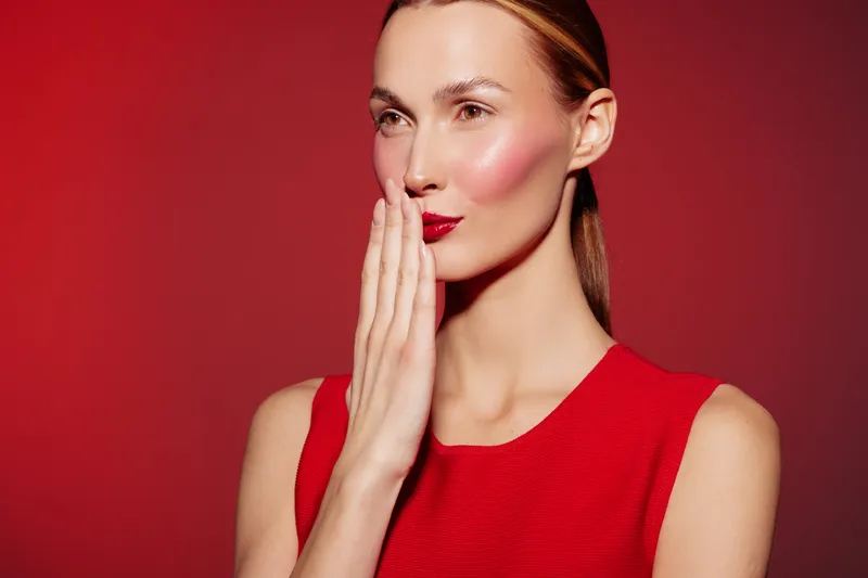 Young woman in red dress with red lipstick covers mouth with hand, posing on red background. Concept of beauty, fashion, makeup, and elegance in studio lighting.