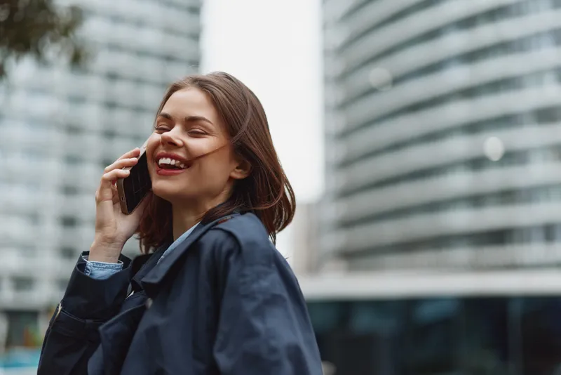 A smiling woman on a smartphone speaks in an urban street setting, capturing a candid moment of everyday communication and connection in a busy city with glass towers.