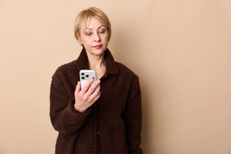 Older woman portrait indoors, holding smartphone with eyes gently closed, wearing dark coat against beige wall, calm moment, daily communication and technology