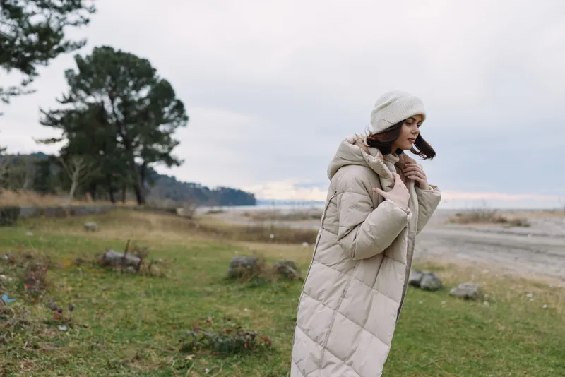 woman, coat, outdoor, wind, landscape, fashion, winter, scenery, A lone figure wrapped in a thick puffer coat and knit hat walks along a windy shoreline meadow under a muted sky, evoking calm, style