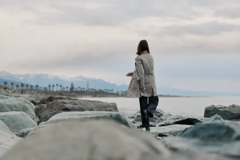 A woman in a coat walks on rocks by the sea with distant mountains and palm trees under an overcast sky. Concept of solitude, nature, and travel exploration.