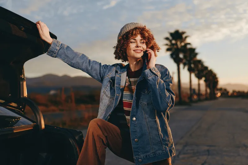 Smiling woman lifestyle outdoors near car at sunset, wearing denim jacket and curly hair, standing on street with warm evening light, relaxed travel pose and sunlit landscape.