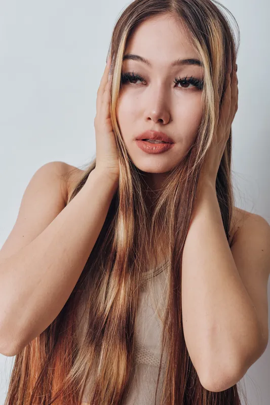 Young woman with long hair touching her head with both hands, natural makeup, and neutral background. Portrait of beauty, fashion, and healthy hair care.