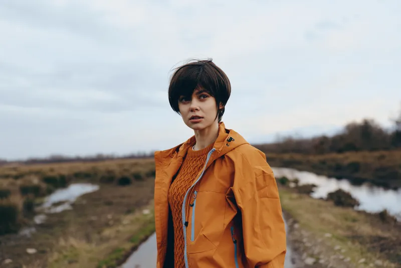 Woman outdoors lifestyle portrait, standing on a rural trail, wearing an orange jacket, short dark hair, calm face, cloudy sky, soft daylight, natural landscape background