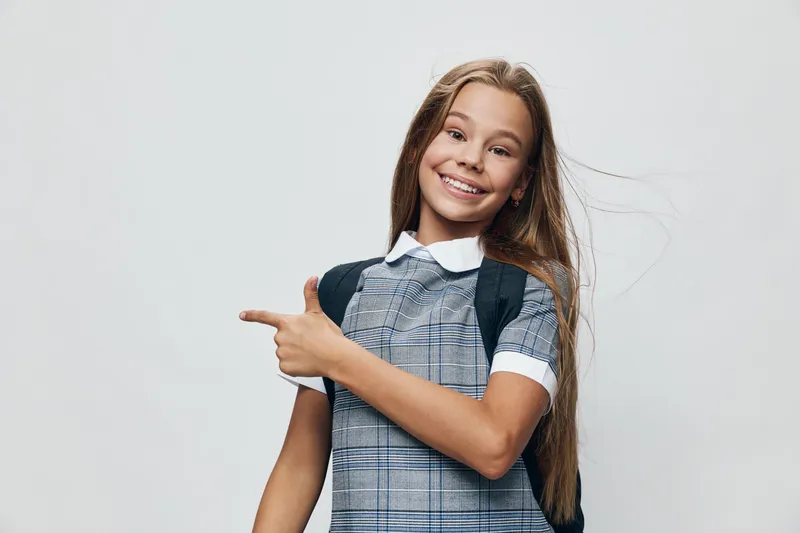 Smiling schoolgirl with long hair wearing checkered dress and backpack pointing with finger on light background. Happy child, education, casual and cheerful girl with positive pose.