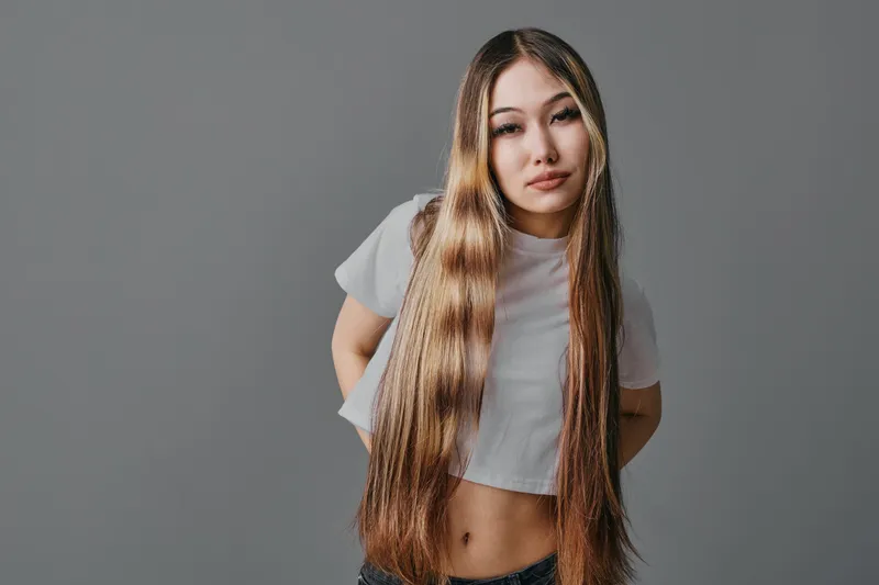 Young woman with long straight hair wearing light crop top standing against gray background looking at camera with calm expression and natural makeup.