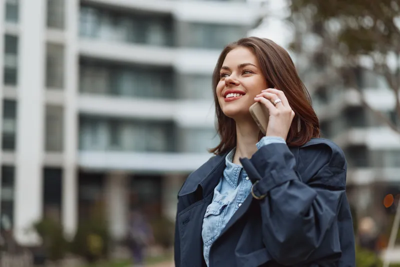 A young woman wearing a denim jacket talks on her phone on a busy city street, smiling as modern buildings rise behind her.