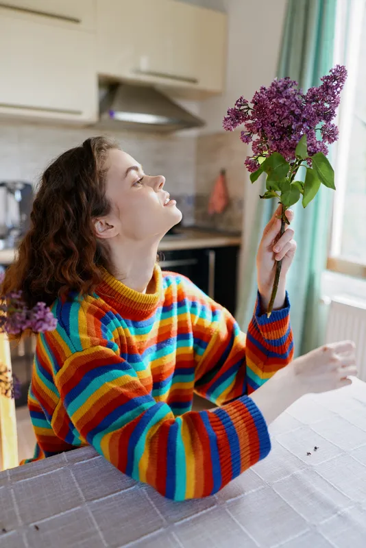 Cheerful woman indoors, smiling with bouquet in hand, colorful striped sweater, sunlit kitchen, cozy moment, home lifestyle, natural light, relaxed pose, fresh flowers, everyday joy