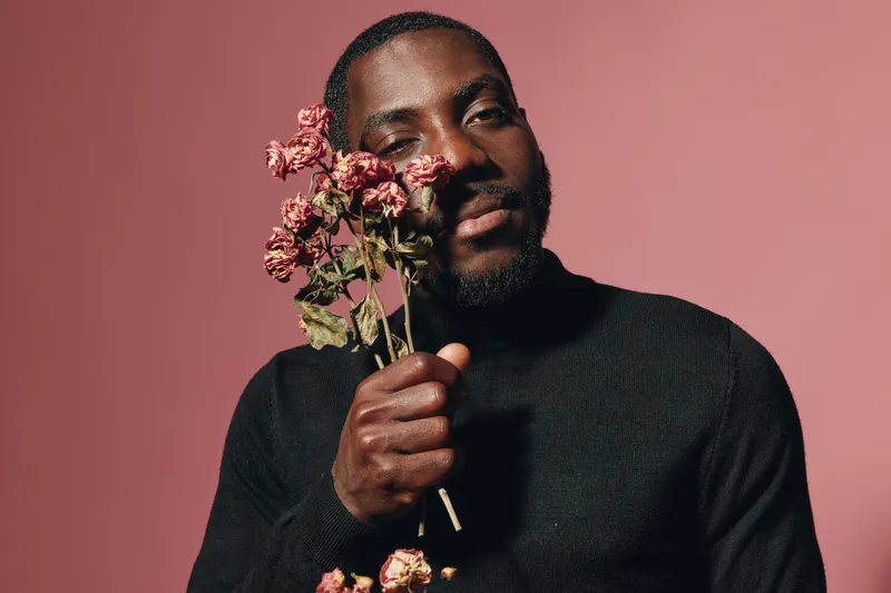 African man with dark skin holding dried flowers close to face in creative conceptual visual style. Portrait with pink background and black sweater for artistic expression and emotion.