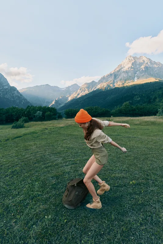 Young woman with long hair wearing an orange beanie jumps playfully on a backpack in a green meadow surrounded by mountains under a clear sky.