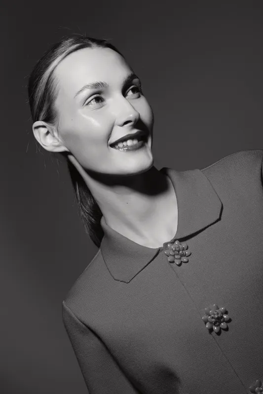 Smiling Caucasian woman with fair skin and sleek hair wears textured coat with decorative buttons. Studio portrait in black and white with soft lighting highlights facial features.