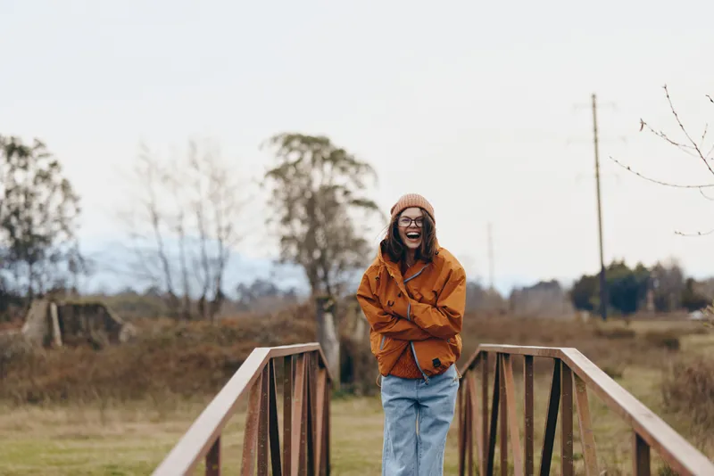 Woman outdoors, laughing in a calm rural field, wearing knit hat and orange jacket, arms crossed by a wooden fence, lifestyle moment, soft daylight, natural colors